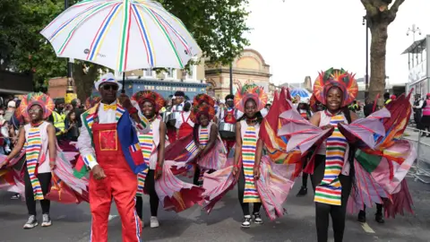 PA Media Performers in colourful, coordinated costumes dance in the street during Notting Hill Carnival, with one man holding a large striped umbrella leading a group of smiling girls wearing vibrant headpieces and wing-like fabric.