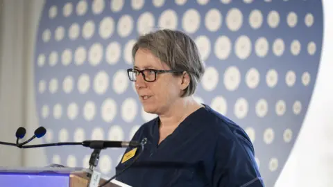 A woman with short grey hair wearing dark blue medical scrubs stands in front of a podium and microphone.