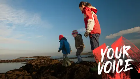 Three children walking on rocks surrounded by sea water. The sky is blue but they are wrapped up against the cold. 
