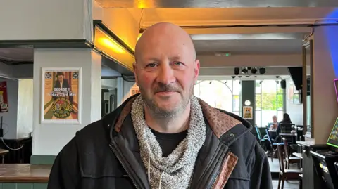 Holly Nichols/BBC A man with a bald head and wearing a coat is standing in a pub and smiling as he looks into the camera