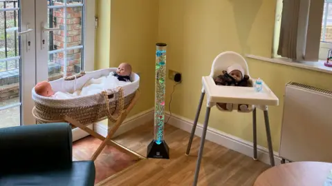 A room with a basinet in it. There is a baby doll sat at each end of it. On the right of the picture there is a high-chair with a doll sat in it.