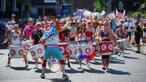A shot looking down a large pride parade - in the foreground are a samba band, all holding red and white drums and dressed for a summer's day with rainbow accessories. The leader has his back to the camera to face and instruct the band, and wears a pair of rainbow wings