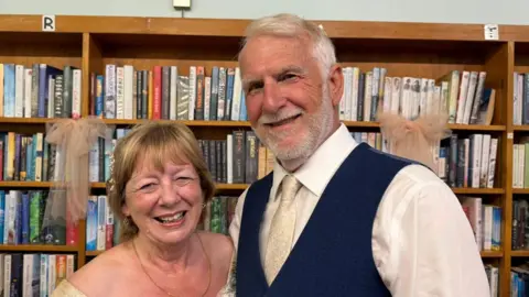 The photo shows two people smiling at the camera. The woman on the left is wearing a cream-coloured dress. The man on the right is wearing a navy waistcoat, cream tie, and white shirt. They're standing in front of shelves that are filled with books, and have two pink bows hanging from them. 