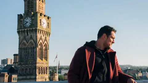 BBC/Magical Society A man in a red jacket leans on a rail and looks away from the camera. A huge clock tower stands in the background.