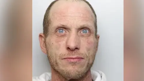 A mugshot of a man who is wearing a white jumper. He has blonde facial hair and short, shaven hair which reveals a receding hairline.