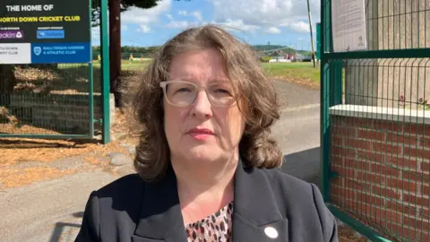 A woman with long brown hair wears brown-framed glasses. She has a black suit with a cheetah print top underneath. She looks into the camera wearing red lipstick. Behind her is two large green metal gates which are open, connected to a low red-brick wall. Behind the wall are green cricket pitches. 