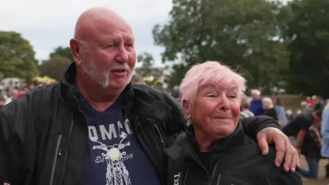 A man with his arm around a woman. The man is bald with grey facial hair. He is wearing a blue T-shirt with a white image of a motorcycle on and a black jacket. The woman has light pink hair and is wearing a black jacket. They are stood in a field. 