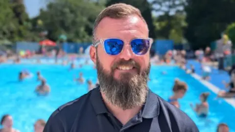 A man with short brown hair and mirrored sunglasses with a dark blue shirt and a blurred outdoor pool behind him