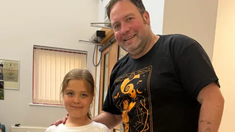 AILSA'S AIM The picture shows the top half of a man on the right of the image smiling looking at the camera. He has his hands on the shoulder of a girl who is looking at the camera and you can see head and shoulders of. They are standing in a corridor with white walls. Behind them you can see a light brown door with a window in it, a window covered by a blind and part of a notice board.
