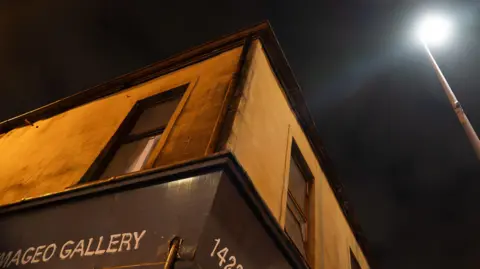 A photo of the corner of a building, taken from street level, looking up at two first floor windows. It is night time.