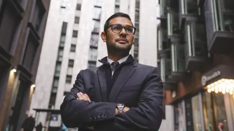 Penguin PR Dan Miller with his arms folded and looking off camera into the distance. He's standing in a city with tall buildings around him.