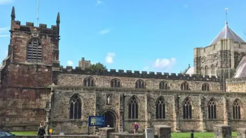 An exterior view of Bangor Cathedral, it is a clear day and there are two people in the foreground of the picture, one walking towards the front door of the cathedral.