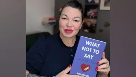 Handout Kiki Deville smiles at the camera holding her book, which has a purple cover and is illustrated with a love-heart affixed with a sticking plaster