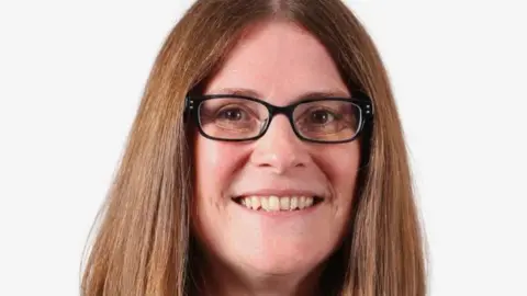 A woman with long brown hair and dark framed glasses smiles in front of a white background