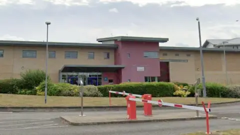 Google Exterior of the prison, a red and cream wide building, with red and white barriers in front of it.
