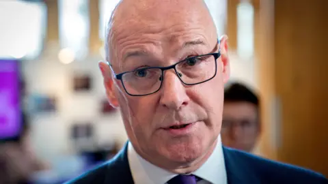 John Swinney - a bald man with glasses and wearing a suit - speaks to media in a lobby