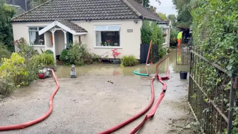 A bungalow is surrounded by several feet of water. Three fire hoses trail along the ground to pump out the water.