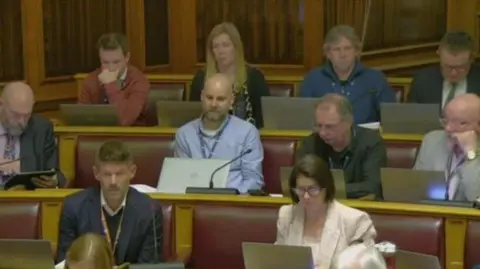 Dorset Council Councillors in the chamber at Dorset Council. Ten councillors - eight men and two women are sitting across three rows of seats. Each is looking at an open laptop.