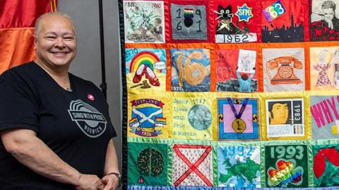Stevie Savage stands next to Norwich Pride patchwork quilt created by the Sew Gay stitchers