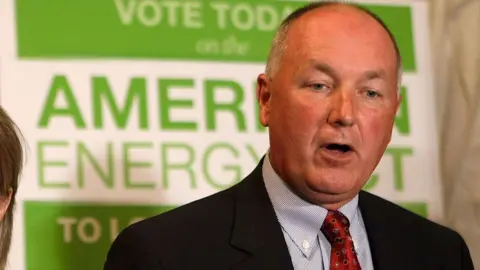 Pete Hoekstra, with short grey hair, and in a dark suit coat, red tie and patterned blue shirt, stands in from of a green and white campaign sign promoting American energy. 