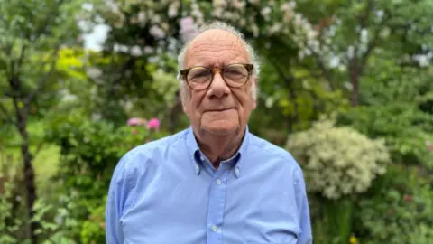 A man with thin grey hair and glasses in a long-sleeved blue shirt in front of green bushes