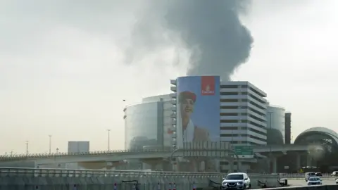 Reuters Black smoke rising from a building in Dubai. There is a huge Emirates advert on the side of the building featuring a female member of cabin crew. There are roads and cars in front of the building.
