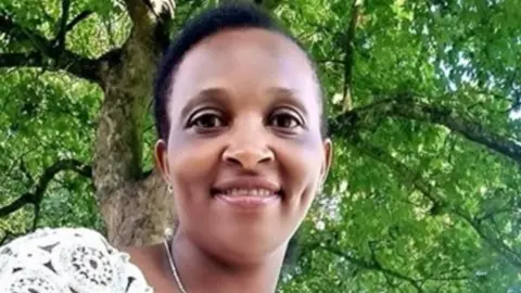 Family Irene is sat outside under a tree and is smiling at the camera. She has a white patterned top on and short, crpopped hair.