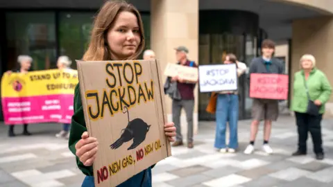 PA Media Environmental campaigners from Friends of the Earth protest outside a UK government building in Edinburgh against the decision to approve Shell's Jackdaw gas field in June 2022. A woman with shoulder-length brown hair is in the foreground holding as placard reading: Stop Jackdaw. No new gas, no new oil. Begins her is a row of campaigners holding placards saying #Stop Jackdaw.