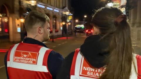 Man and woman wearing orange and white Northampton Guardians jackets in a street at night
