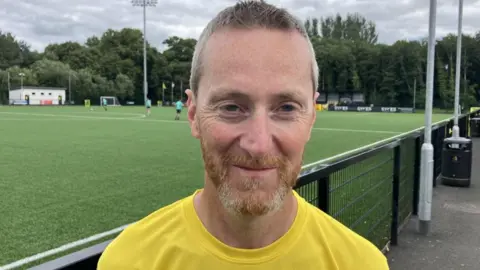 Man with ginger beard wearing yellow T-shirt, standing at the side of a football pitch