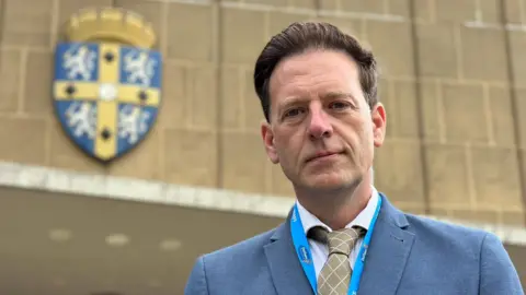 New Durham County Council leader Andrew Husband outside County Hall. He is wearing a light blue suit jacket, a white shirt and a gold tie. His brown hair is swept back.
