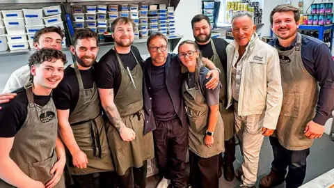 Norman's A group of people pose for a picture with actor Stephen Graham and Bruce Springsteen. Most of them are wearing brown aprons as they stand in a kitchen area.