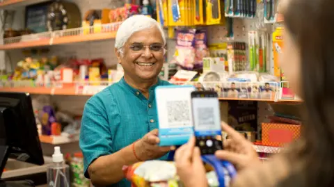 Getty Images Female customer (blurred) scanning QR code with smartphone and paying to a senior male owner at checkout counter in a supermarket. 