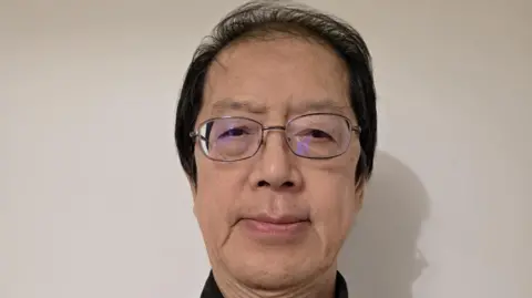 This is a head shot of Joe Wong who is looking at the camera. He is standing in front a white wall.