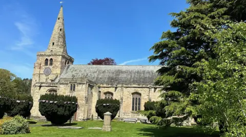 The church. Is is a large building made from yellow stone with a spire at one end, and trees and bushes in front of it.