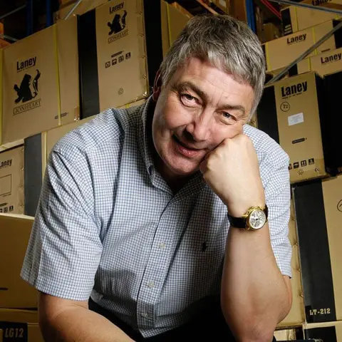 Getty Images A smiling Lyndon Laney wears a checked short-sleeved shirt and is sitting in front of a pile of boxes 