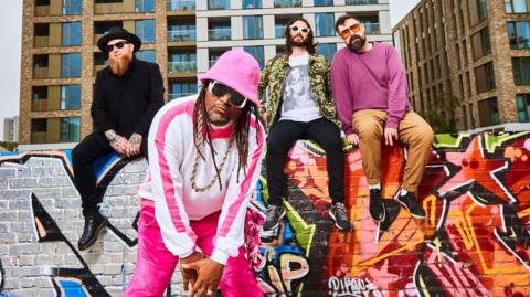 Skindred in very bright clothing posing against a graffitied wall
