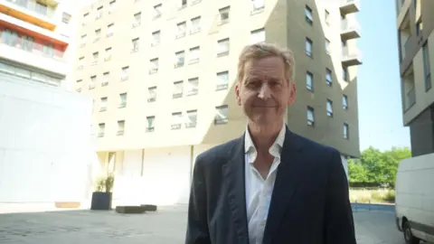 John Fairhall/BBC John Howard wearing a suit standing in front of a block of flats while looking directly into the camera