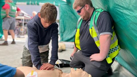 A boy leans over a dummy that is on the ground as he gives chest compressions while a Community First Responder knees next to him