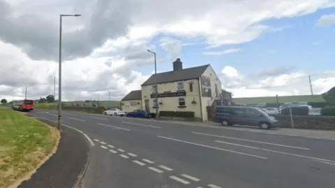 A Street View image of the road which runs past a pub called The Travellers Inn. Several cars can be seen in the pub's car park and a red bus can be seen driving on the road.