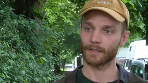 Callum is wearing a black over-shirt and has a beard. He has on a yellow cap. There are vans parked up behind him.