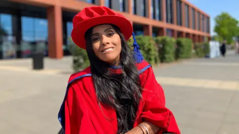 A woman with long dark hair in a red gown and cap smiles at the camera. She is outside a two storey building