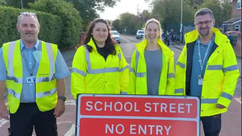 Reading Borough Council Two women and two men standing on a residential road wearing high-vis yellow jackets. One of the men is wearing a whistle around his neck. In front of them is a red and white sign which reads "school street no entry". There are some young children wearing backpacks in the background.