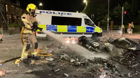 Tom Ingall/BBC A firefighter extinguishes flames from the rubble of a bonfire, pursuing  the riots successful  Harehills, Leeds. A constabulary  van stands crossed  the roadworthy  successful  the background