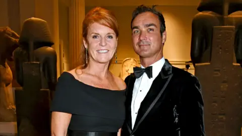 Getty Images Sarah Ferguson in black dress with her hair up stands next to Manuel Fernandez who wears a black velvet jacket, white shirt and bow tie. They are both smiling at the camera. 