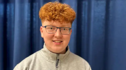 James has red hair. He is wearing glasses and a grey zipped top. 