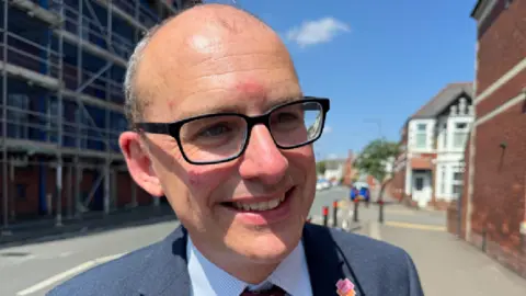 Joseph smiling looking at the reporter who is out of shot. Behind him is a residential street and scaffolding. Joseph is wearing black glasses, a navy suit and a tie.