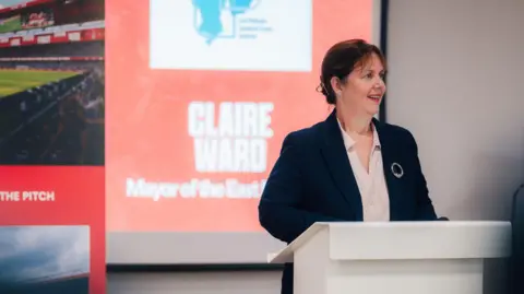 East Midlands Combined County Authority East Midlands mayor Claire Ward speaking at an event at the City Ground. She is wearing a dark blue suit with a light pink shirt and silver brooch. Her brown her is tied back in a hair clip. 