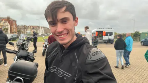 Alaistair Wearing a black hoodie with short dark hair smiling at the camera. There are people loitering in the background in a car park with a few other motorbikes in view.