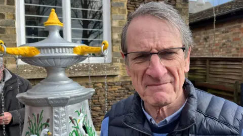 John Devine/BBC Gordon has greyish dark hair swept to the right. He is wearing a black body warmer over a blue and black striped jumper. He is wearing dark framed glasses and the water feature is on the left behind him.